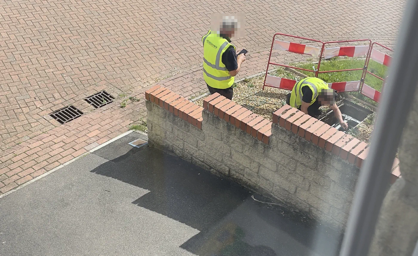 Two engineers in hi-vis vests working at an open BT manhole behind red and white barriers on a brick-paved street