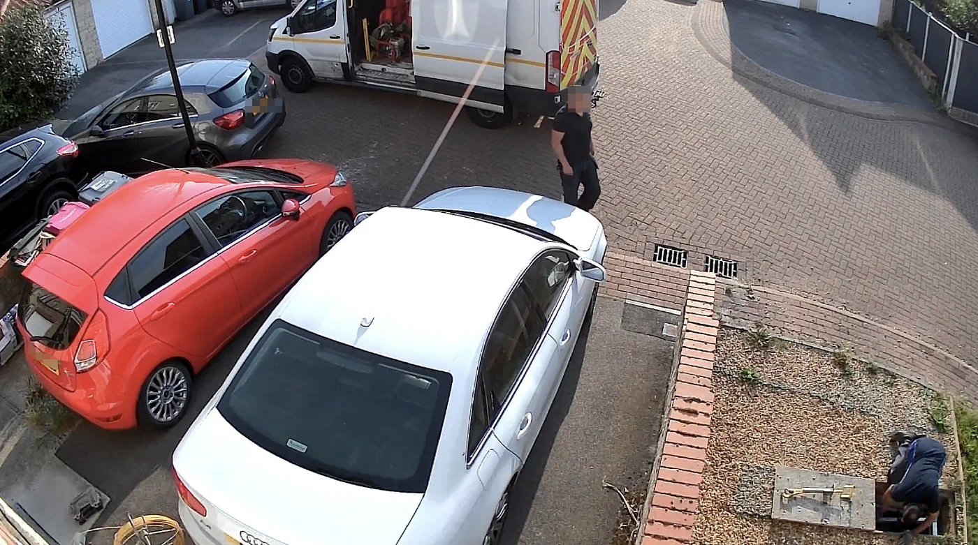 Driveway camera view of a white Virgin Media works van with an engineer kneeling at an open manhole cover in the gravel