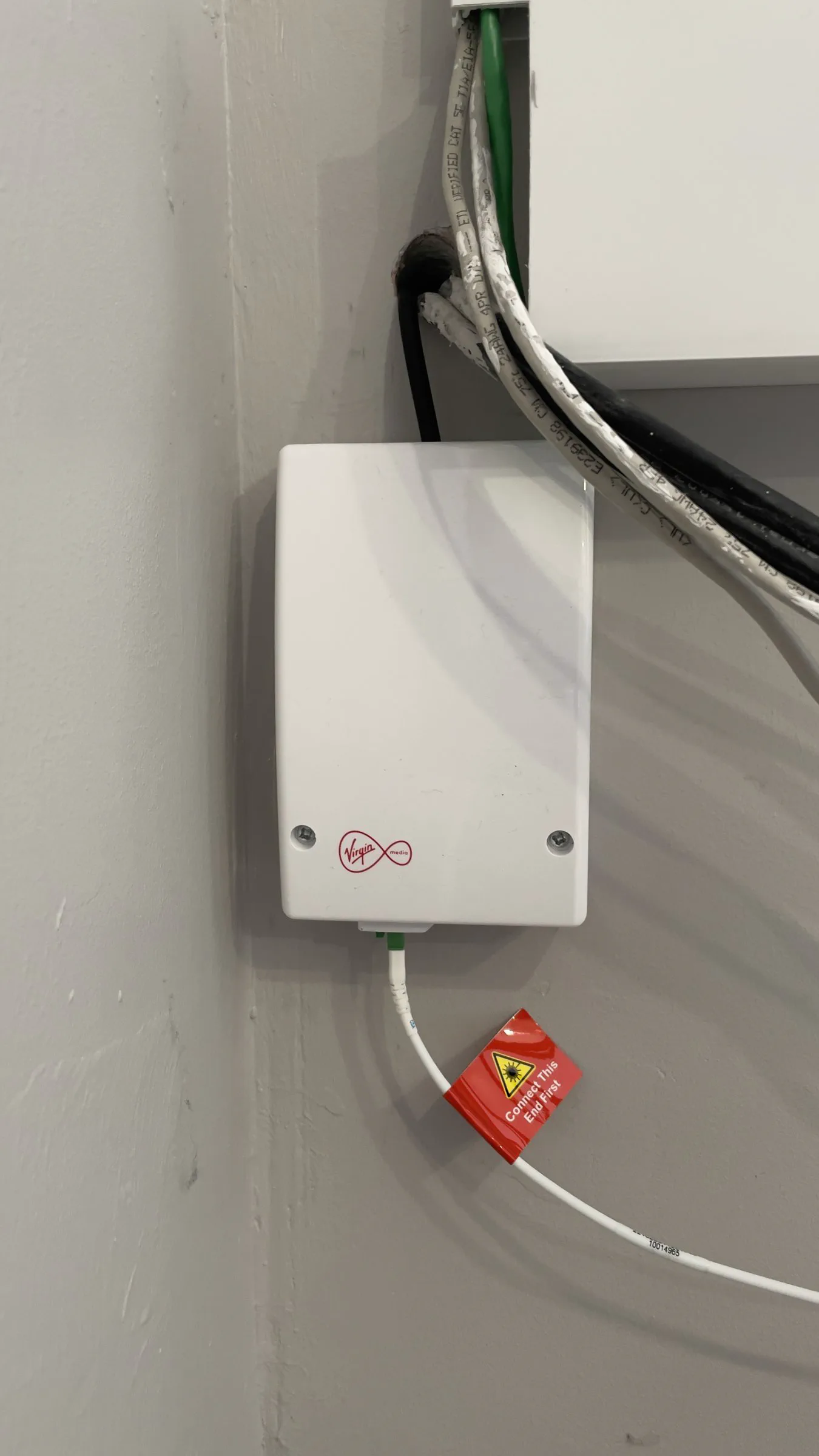 The white Virgin Media indoor fibre box mounted on an internal wall, with the fibre patch lead attached and its red “Connect This End First” tag still on